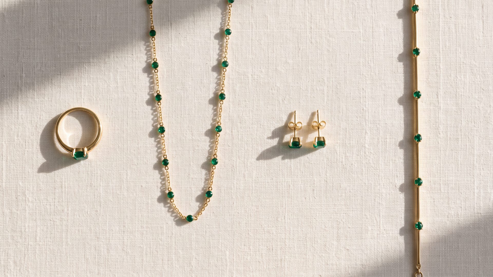 An emerald ring, chain, studs and bracelet laid out in a row on ivory linen