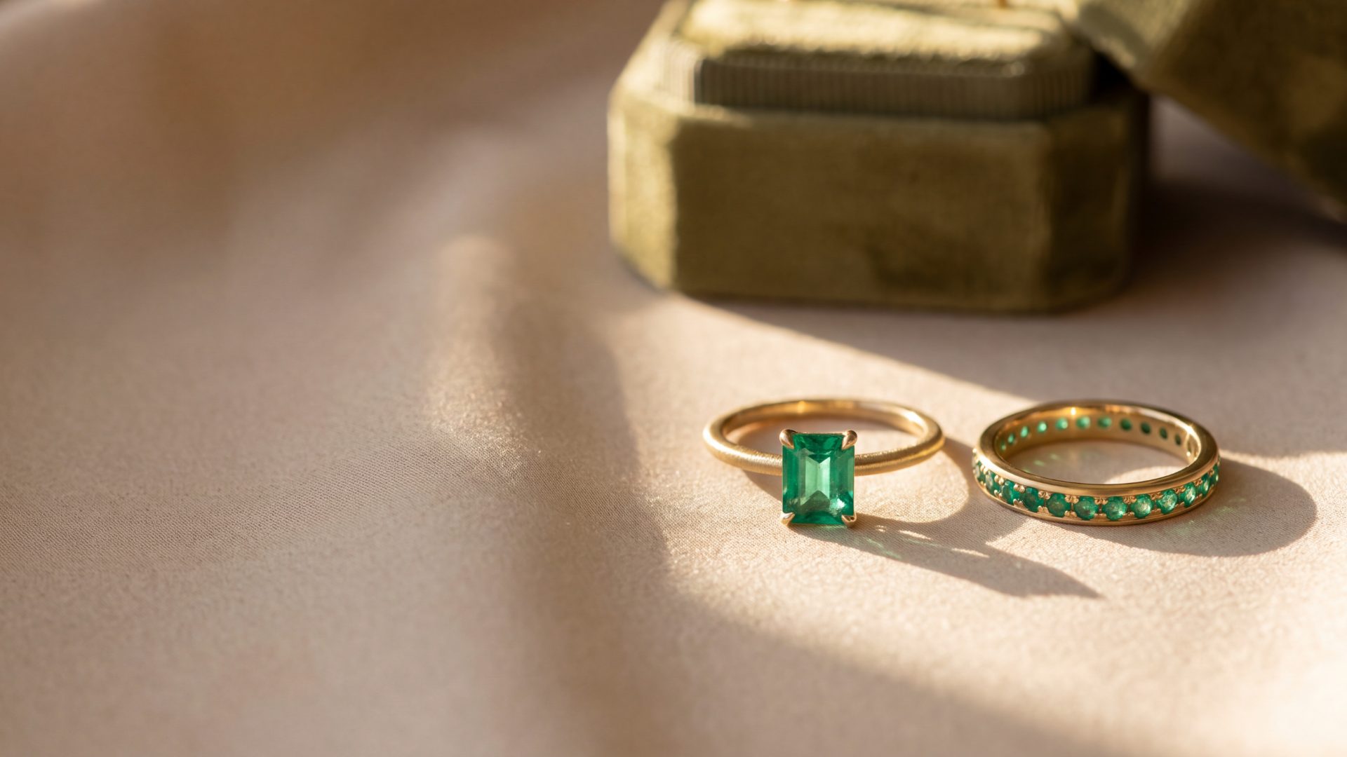 An emerald solitaire ring beside an emerald eternity band on champagne silk in golden-hour light